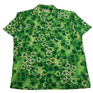 May include: Green short-sleeved shirt with a floral pattern. The shirt has a button-down front, a collar, and a chest pocket. The floral design includes white and orange accents on a green background.