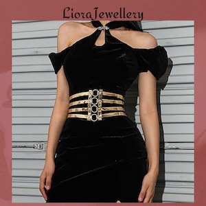 Puede incluir: Un cinturón de metal dorado con cuatro barras horizontales y conectores circulares. El cinturón se lleva sobre un vestido de terciopelo negro. El texto "Liora Jewellery" está en la parte superior.