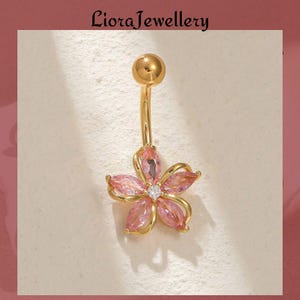 May include: Gold-colored belly button ring with a flower design. The flower has pink petals and a small, clear gem in the center. The jewelry is from Liora Jewellery.