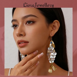 May include: Gold-toned fish-shaped earrings with a blue eye detail and iridescent scales. The earrings have a dangling design with a fan-shaped tail. The brand name "Liora Jewellery" is at the top.