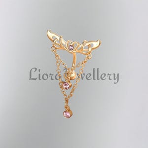 May include: A gold-colored jewelry piece with a decorative design. The piece features a central heart-shaped element and dangling chains with pink gemstones. The text "Liora Jewellery" is visible on the image.