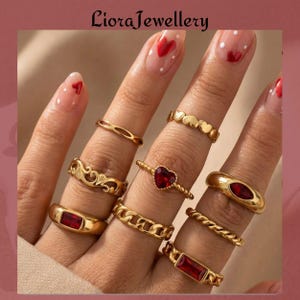 Könnte beinhalten: Eine Auswahl an goldfarbenen Ringen, darunter schlichte Bänder, ein herzförmiger Ring mit rotem Stein und Ringe mit rechteckigen roten Steinen. Die Ringe werden auf Fingern mit Herz-Nagelkunst präsentiert. Der Markenname "LioraJewellery" ist oben zu sehen.