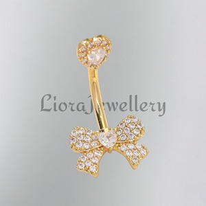 May include: A gold-colored belly button ring with a heart-shaped top and a bow-shaped bottom, both encrusted with clear, sparkling crystals. The text "Liora Jewellery" is visible in the background.