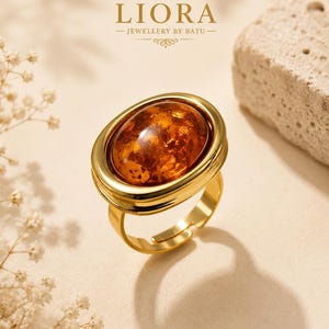 Peut inclure: Une bague dorée ornée d'une grande pierre ovale translucide de couleur ambre, sertie dans un chaton en métal lisse et poli. La bague présente un anneau ouvert réglable. Au-dessus, le texte indique LIORA JEWELLERY BY BATU.