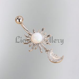 Könnte beinhalten: Ein silberner Bauchnabelpiercing mit einem Sonnen- und Mond-Design. Die Sonne hat strahlende Strahlen und einen zentralen Stein in Opaloptik. Ein Mondsichel-Anhänger ist mit kleinen, klaren Steinen verziert. Der Text "Lion Gallery" ist sichtbar.