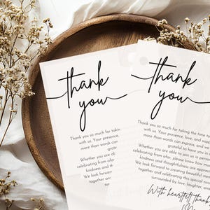 May include: Two cream-coloured thank you cards with elegant black script. The cards feature the words "thank you" in a flowing font. The cards are on a wooden tray with dried flowers, creating a simple, elegant design.