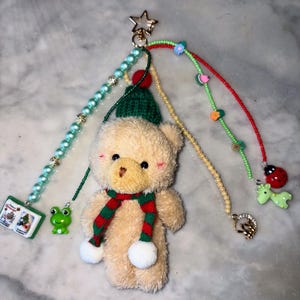 Könnte beinhalten: Ein Schlüsselanhänger mit einem beigefarbenen Teddybären, der eine grüne Strickmütze und einen rot-grün gestreiften Schal trägt. Der Schlüsselanhänger hat mehrere Stränge mit Perlen und Anhängern, darunter ein Frosch, ein Buch, ein Marienkäfer und ein Herz.