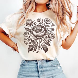 May include: Cream-colored t-shirt featuring a detailed black and white floral design. The graphic includes a large peony surrounded by smaller flowers and leaves. The shirt has short sleeves and a relaxed fit, suitable for casual wear.