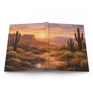 May include: Hardcover journal featuring a desert landscape with cacti, a dirt path, and a sunset. The cover art showcases a vibrant scene with warm colors, including orange, yellow, and purple hues. The journal is open, revealing the interior pages.