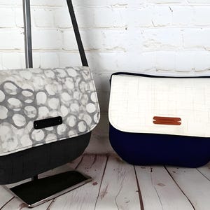 Puede incluir: Dos bolsos tipo mensajero. Uno tiene un estampado gris y blanco en la solapa y una base negra. El otro tiene una solapa blanca y una base azul marino. Ambos bolsos tienen una correa y un pequeño acento rectangular.