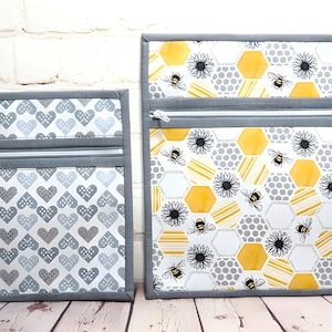 Op de afbeelding: Twee zakjes met ritssluiting. Eén heeft een patroon van grijze en witte harten. De andere heeft een honingraatontwerp met gele, witte en grijze zeshoeken, bijen en bloemen. Beide hebben een grijze rand.