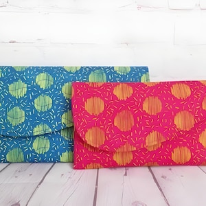 Puede incluir: Dos bolsos de mano de tela. Uno es azul con patrones circulares verdes y blancos, y el otro es rosa con patrones circulares naranjas y amarillos. Los bolsos son rectangulares con un cierre de solapa curvada.