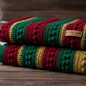 May include: Two folded, handmade crochet blankets in red, green, and gold. The blankets have a textured, bobble stitch design and are stacked on top of each other. A small leather tag with text is attached to the top blanket.