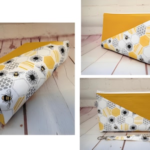 May include: A yellow and white zippered pouch with a bee and honeycomb pattern. The pouch features a diagonal yellow panel and a matching wrist strap. The design includes black bees and flowers on a white background.