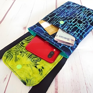 Puede incluir: Un bolso bandolera para teléfono hecho a mano con una correa negra. El bolso contiene un teléfono rojo, tarjetas de crédito y un papel doblado. El bolso tiene una parte superior con estampado azul y negro y una parte inferior con estampado amarillo y verde.