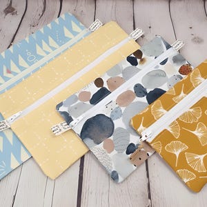 May include: Five rectangular zippered pouches in various patterns and colors. Designs include blue and white geometric shapes, yellow with white dots, gray and blue watercolor stones, and a mustard yellow ginkgo leaf pattern. Each pouch has a white zipper.