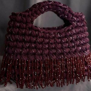 Può includere: Una borsa bordeaux con manico intrecciato e frangia di perline. La borsa è realizzata in un tessuto strutturato con fili d'argento intrecciati. La frangia è composta da piccole perline rosso scuro.