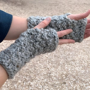 Puede incluir: Un par de guantes sin dedos grises, tejidos a mano con un hilo grueso. Los guantes son de color gris claro con motas de gris más oscuro y están diseñados para cubrir la mano hasta los nudillos. Un accesorio acogedor para el clima fresco.