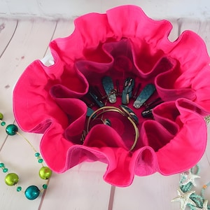 May include: A bright pink, ruffled jewelry organizer with multiple compartments. The organizer is filled with jewelry, including bracelets and lipsticks. Two necklaces with green and teal beads are on either side.