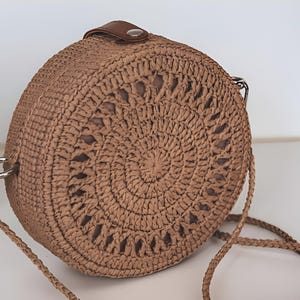 May include: A round, woven straw handbag with a brown leather strap and silver-tone hardware. The bag features a circular, open-weave design on the front. The shoulder strap is made of braided straw. The bag is tan in color.