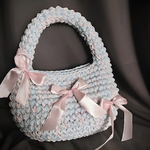 May include: A handmade crochet handbag in light blue and white, adorned with pink satin bows. The bag features a rounded handle and is designed with a textured stitch pattern. The bag is a soft, pastel-colored accessory.