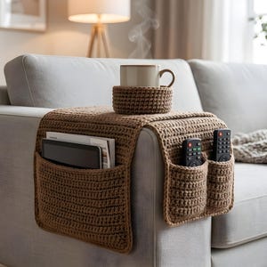 Puede incluir: Un organizador de reposabrazos de sofá marrón tejido a ganchillo con bolsillos para mandos a distancia, una tableta y revistas. Un posavasos a juego sostiene una taza beige con vapor saliendo. El organizador está en un sofá gris claro.