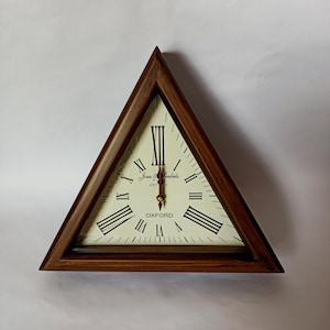 May include: Triangular wooden wall clock with a cream-colored face and black Roman numerals. The clock has a dark brown wooden frame and red clock hands. The text "James & Hendricks EST 13" and "OXFORD" are printed on the clock face.