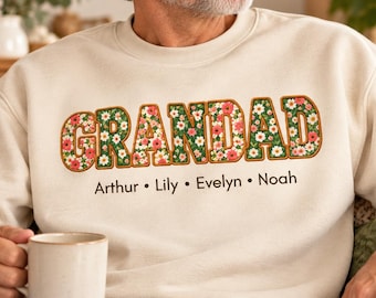 May include: A beige crewneck sweatshirt featuring the word GRANDAD in large block letters filled with a colorful floral pattern of red, pink, and white flowers on a green background. Below the text are the names Arthur, Lily, Evelyn, and Noah.