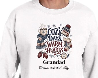 May include: A white crewneck sweatshirt featuring a graphic print with the text "COZY DAYS, WARM HEARTS", "Grandad", and "Emma, Noah & Lily". The design includes illustrations of a mug, a knit hat, a scarf, socks, mittens, and a teapot.