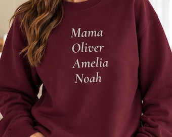 May include: A maroon crewneck sweatshirt featuring white text printed on the chest. The text is stacked vertically and reads: Mama, Oliver, Amelia, Noah. The sweatshirt has long sleeves with ribbed cuffs and a ribbed neckline.
