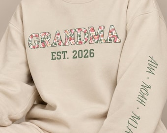 May include: A beige crewneck sweatshirt featuring the word GRANDMA in a floral pattern with pink and green flowers. Below, the text EST. 2026 is printed in dark green. The left sleeve displays the names AVA, NOAH, and MIA in dark green text.