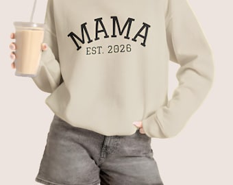 May include: A beige long-sleeve crewneck sweatshirt featuring the black text MAMA arched over the text EST. 2026 in a serif font. The sweatshirt is paired with gray denim shorts, and a clear plastic tumbler with a straw is held in one hand.