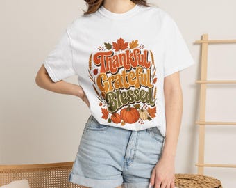 May include: A white short-sleeve t-shirt featuring a graphic print with the text Thankful Grateful Blessed in orange, yellow, and green. The design includes illustrations of autumn leaves, wheat stalks, and three orange pumpkins.