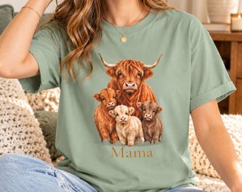 May include: A sage green short-sleeve t-shirt featuring a graphic print of a large Highland cow standing behind three smaller Highland calves. The word "Mama" is printed in a simple, orange-toned font directly below the animal illustration.