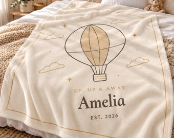 May include: A cream-colored fleece blanket featuring a central graphic of a hot air balloon surrounded by small clouds and stars. The text reads UP, UP & AWAY, the name Amelia, and EST. 2026 in dark brown lettering with a thin gold border.