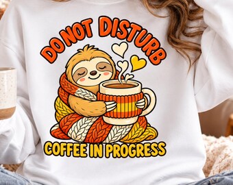 May include: A white sweatshirt featuring a graphic of a sloth wrapped in a thick orange, yellow, and white knit blanket, holding a coffee mug. The text reads DO NOT DISTURB above and COFFEE IN PROGRESS below, with small heart shapes floating above the mug.
