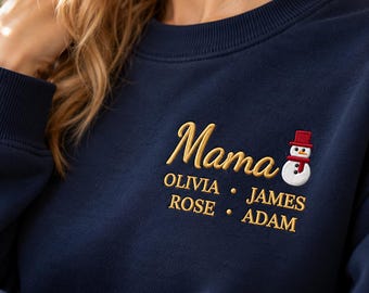 May include: A navy blue crewneck sweatshirt featuring gold embroidered text that reads Mama, followed by the names OLIVIA, JAMES, ROSE, and ADAM. To the right of the word Mama is a small embroidered snowman wearing a red hat and scarf.