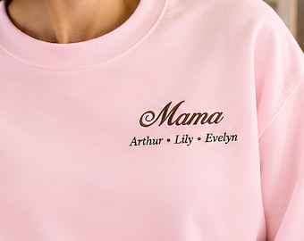 May include: A light pink crewneck sweatshirt featuring dark brown embroidery on the chest. The text reads "Mama" in a large script font, followed by the names "Arthur • Lily • Evelyn" in a smaller, clean serif font centered below it.