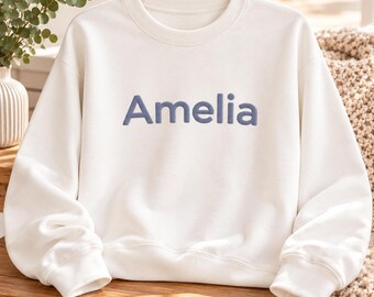 May include: A white crewneck sweatshirt featuring the name Amelia printed in a raised, matte blue font across the center of the chest. The garment has long sleeves with ribbed cuffs and a ribbed neckline, providing a clean and simple appearance.
