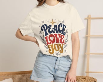 May include: A cream-colored short-sleeve t-shirt featuring the text PEACE LOVE JOY in navy blue, red, and gold. The design includes a star, snowflakes, and pine branch illustrations surrounding the words in a centered graphic print.