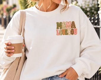 May include: A white crewneck sweatshirt featuring the text MAMA LOVE YOU printed on the left chest. The letters are filled with various patterns including green, pink gingham, floral, and leopard print. The sweatshirt has long sleeves and a ribbed collar.