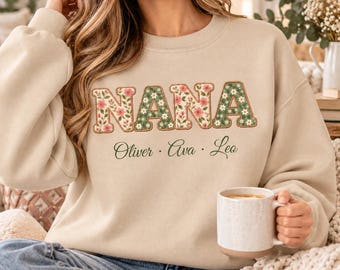May include: A beige crewneck sweatshirt featuring the word NANA in large, floral-patterned block letters with pink and white flowers on a green background. Below the word, the names Oliver, Ava, and Leo are printed in a dark green script font.