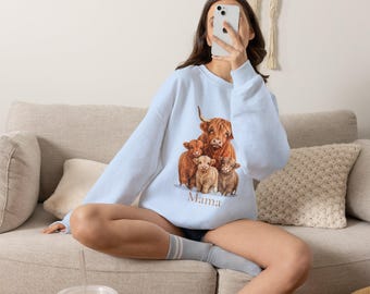 May include: A light blue crewneck sweatshirt featuring a graphic print of a group of brown Highland cattle with the word "Mama" printed in brown text underneath the animals.