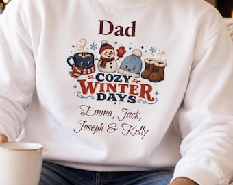 May include: A white crewneck sweatshirt featuring a graphic print with the text Dad, Cozy Winter Days, and the names Emma, Jack, Joseph & Kelly. The design includes a steaming mug, a snowman, a blue knit hat, and brown winter boots.