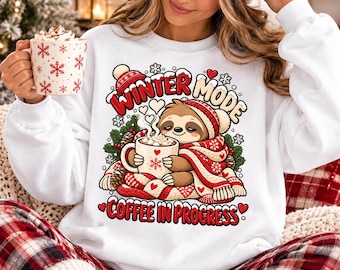 May include: A white long-sleeve sweatshirt featuring a graphic of a sloth wearing a red and white knit hat and scarf, holding a mug of coffee. The text reads "WINTER MODE" and "COFFEE IN PROGRESS" in red letters, surrounded by snowflakes and pine.