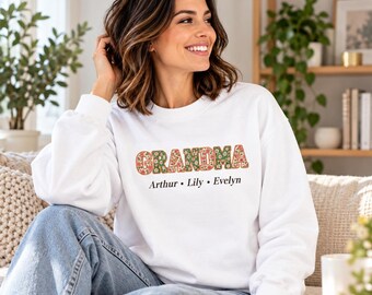 May include: A white long-sleeve crewneck sweatshirt featuring the word GRANDMA printed in a floral pattern with red, pink, and green flowers. Below the word, the names Arthur, Lily, and Evelyn are printed in a simple black sans-serif font.