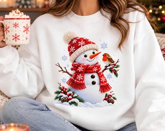 May include: A white crewneck sweatshirt featuring a graphic of a snowman wearing a red knit hat with a pom-pom and a matching red scarf. The snowman has a carrot nose, two black buttons, and is perched next to a small robin and pine branches.