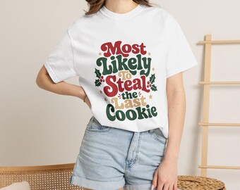 May include: A white short-sleeve t-shirt featuring the red, green, and beige text Most Likely To Steal the Last Cookie, decorated with small green holly leaves, red berries, and gold stars printed on the front center of the garment.