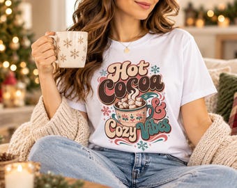 May include: A white short-sleeve t-shirt featuring a graphic print with the text Hot Cocoa & Cozy Vibes, a mug filled with marshmallows, and snowflake accents. The design uses brown, pink, and teal colors on the front of the shirt.
