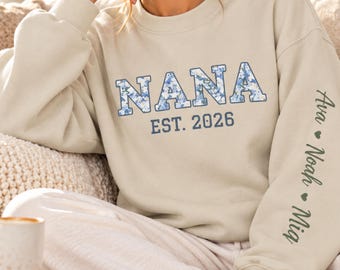 May include: A beige crewneck sweatshirt featuring the word NANA in blue floral block letters with the text EST. 2026 underneath. The left sleeve displays the names Ava, Noah, and Mia in dark green script, separated by small dark green hearts.
